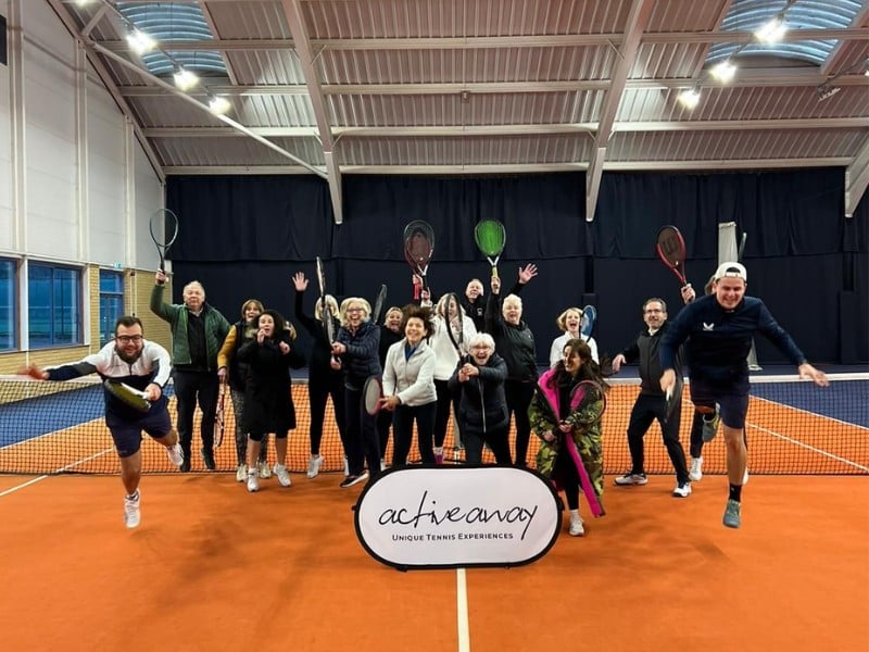 Group of enthusiastic tennis players posing on an indoor court at DL Cheshire Oaks, holding rackets behind an 'Active Away' banner.