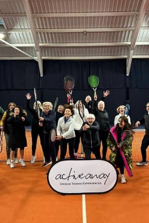 Group of enthusiastic tennis players posing on an indoor court at DL Cheshire Oaks, holding rackets behind an 'Active Away' banner.
