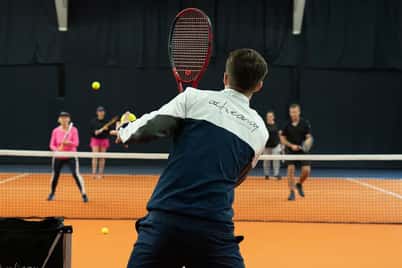 Tennis coach serving on indoor court with players ready for training, showcasing dynamic sports practice.