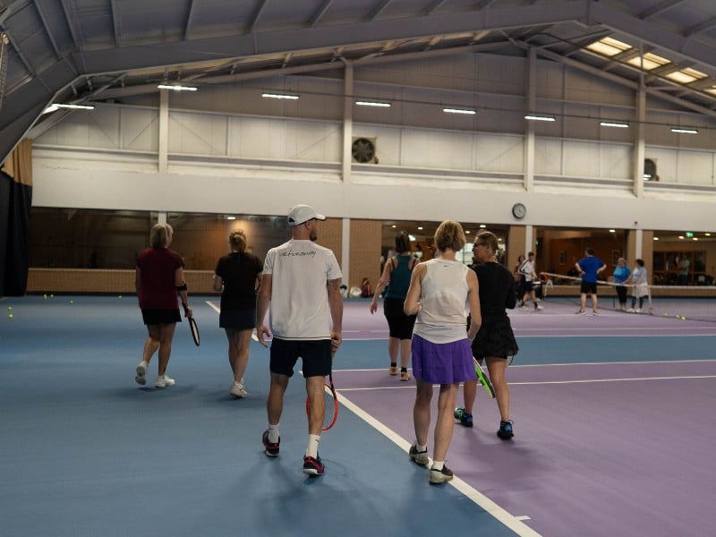 Group of tennis players on an indoor court with blue and purple surfaces, featuring high ceilings and modern lighting.