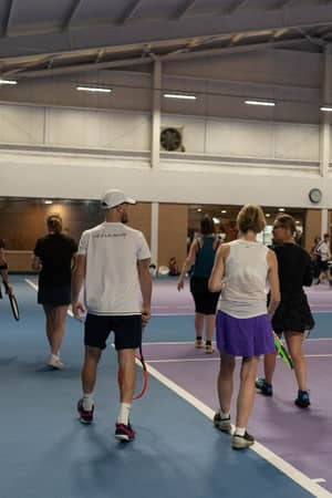 Group of tennis players on an indoor court with blue and purple surfaces, featuring high ceilings and modern lighting.
