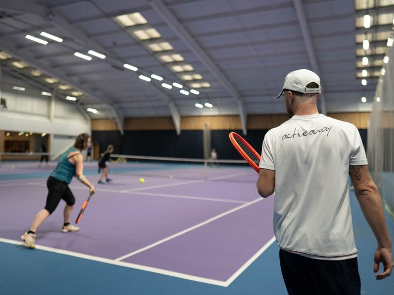 Tennis coach observes two players practising on a purple indoor court at DL Chorley, with vivid lighting and spacious layout.