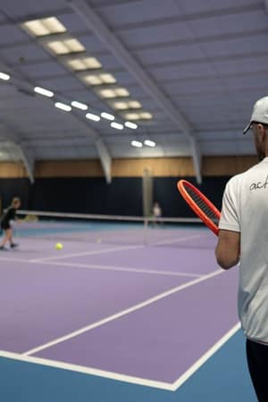 Tennis coach observes two players practising on a purple indoor court at DL Chorley, with vivid lighting and spacious layout.