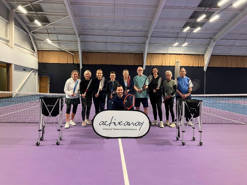 Group photo of ten people holding tennis rackets on an indoor court with an Active Away banner and trolleys.
