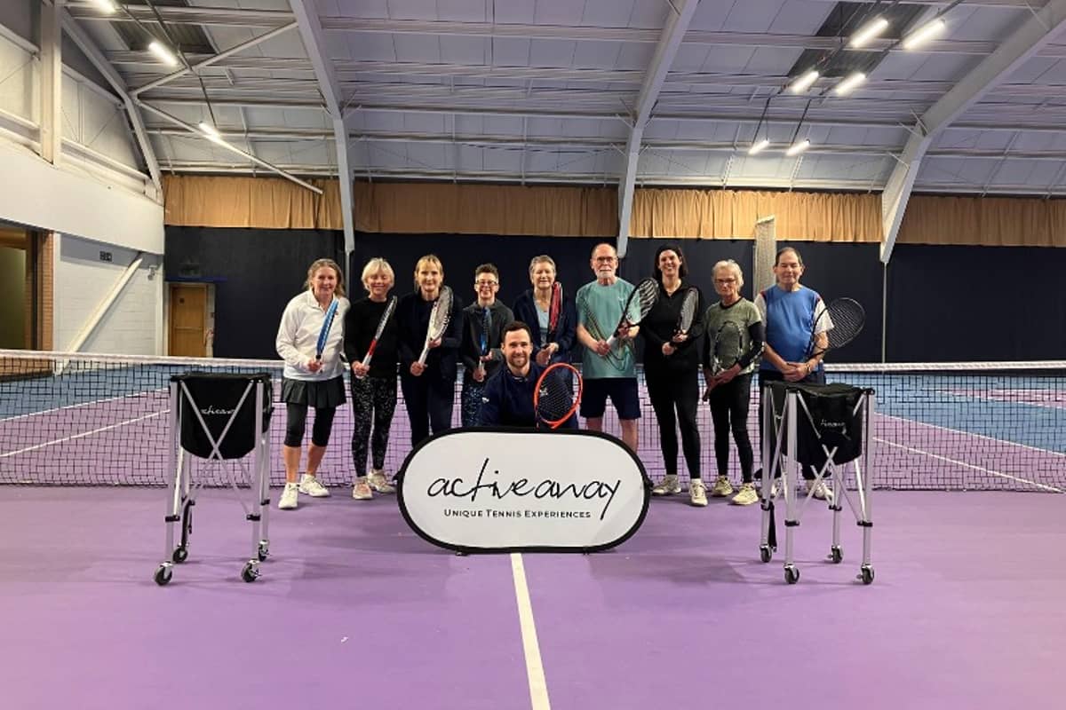 Group photo of ten people holding tennis rackets on an indoor court with an Active Away banner and trolleys.