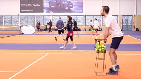 Indoor tennis court at DL Edinburgh with players practising and a coach holding a basket of tennis balls.