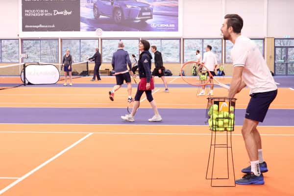 Indoor tennis court at DL Edinburgh with players practising and a coach holding a basket of tennis balls.