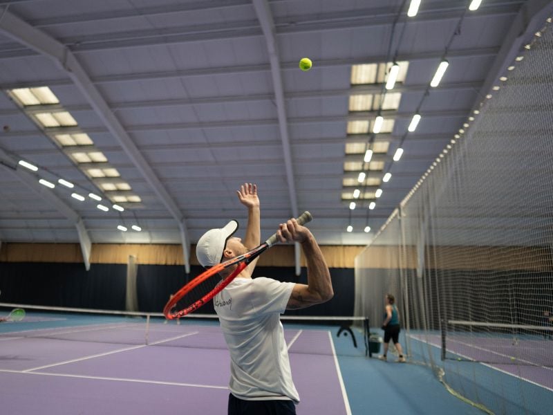 Tennis coach in a white cap serving on a purple indoor court, with bright overhead lights and another player seen in the background.