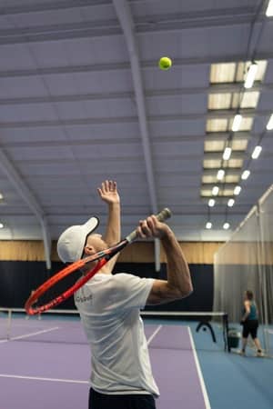 Tennis coach in a white cap serving on a purple indoor court, with bright overhead lights and another player seen in the background.