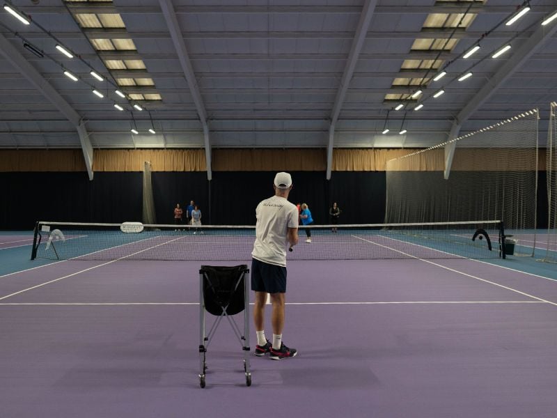 Tennis coach conducting a training session on an indoor court at DL Edinburgh, with players practicing in the background.