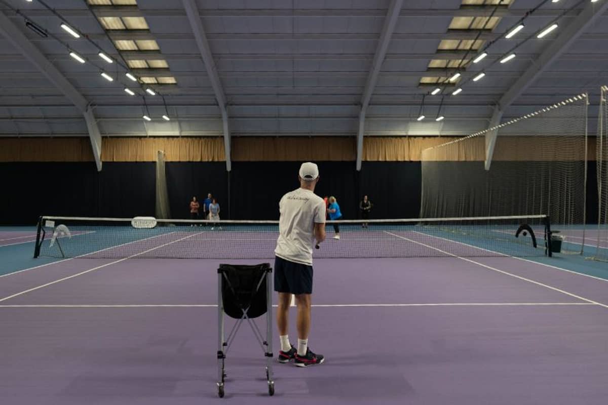 Tennis coach conducting a training session on an indoor court at DL Edinburgh, with players practicing in the background.