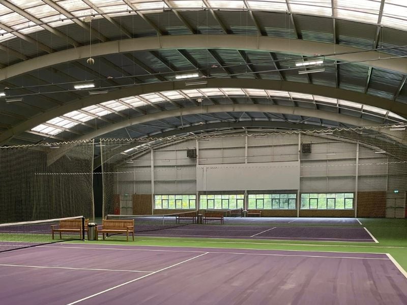 Indoor tennis court at DL Edinburgh with purple and green surface, large roof, natural light, and wooden benches.