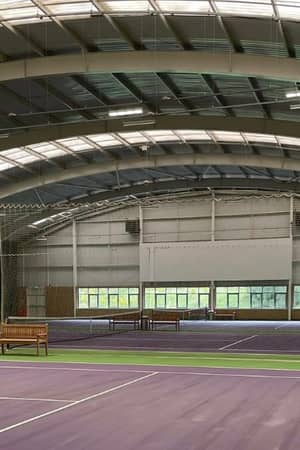 Indoor tennis court at DL Edinburgh with purple and green surface, large roof, natural light, and wooden benches.