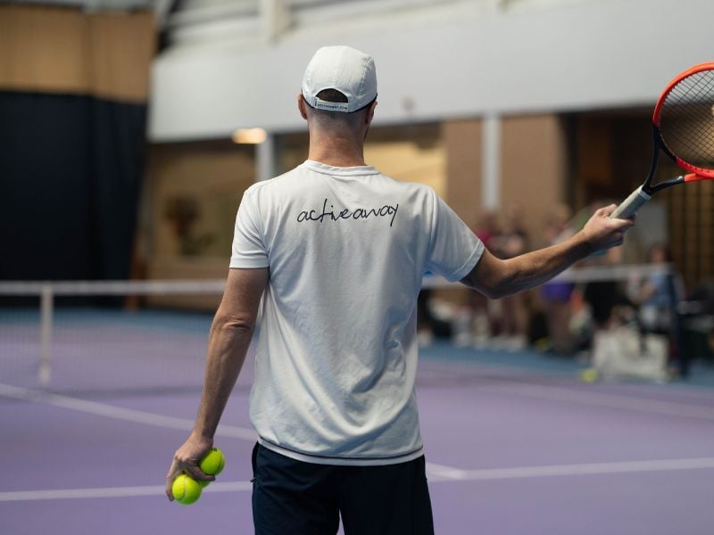 Tennis coach at DL Finchley, wearing a white achieve t-shirt and holding a racquet and tennis balls on an indoor court.