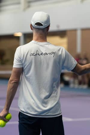 Tennis coach at DL Finchley, wearing a white achieve t-shirt and holding a racquet and tennis balls on an indoor court.