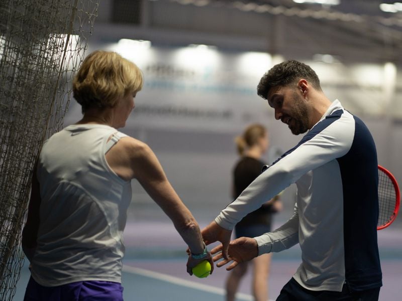 Tennis coach teaching a player at DL Finchley Gallery indoor court, focusing on grip and stance with another player in the background.