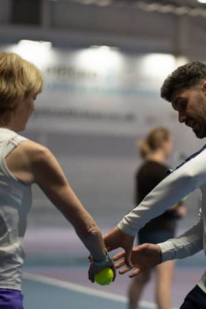 Tennis coach teaching a player at DL Finchley Gallery indoor court, focusing on grip and stance with another player in the background.