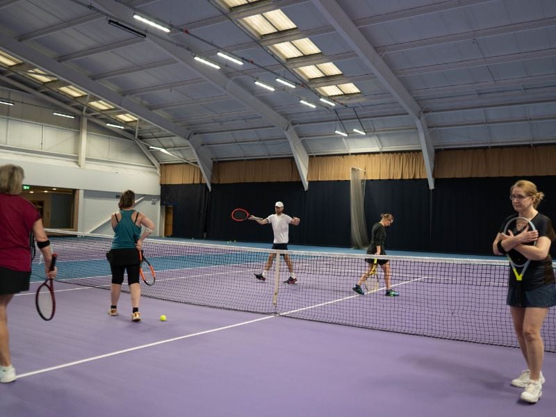 People engaging in a tennis drill session at DL Finchley, holding rackets on an indoor court with a purple floor.