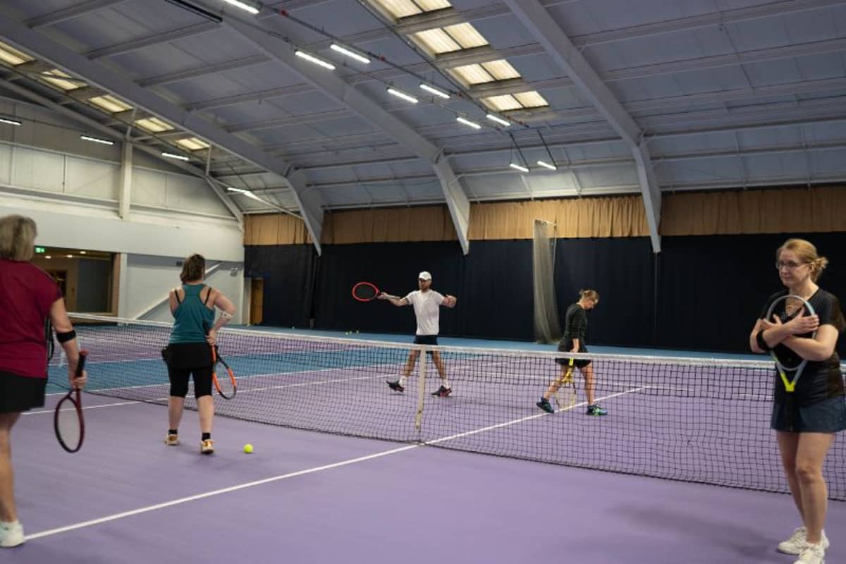 People engaging in a tennis drill session at DL Finchley, holding rackets on an indoor court with a purple floor.