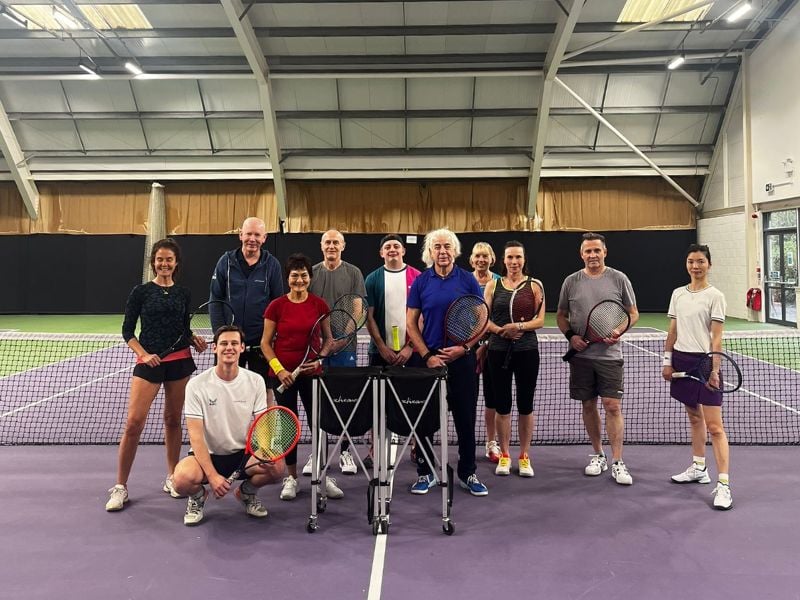 Group of tennis players at DL Finchley Gallery indoor tennis courts, holding rackets, with a steel ceiling and curtains in the background.