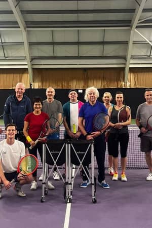 Group of tennis players at DL Finchley Gallery indoor tennis courts, holding rackets, with a steel ceiling and curtains in the background.