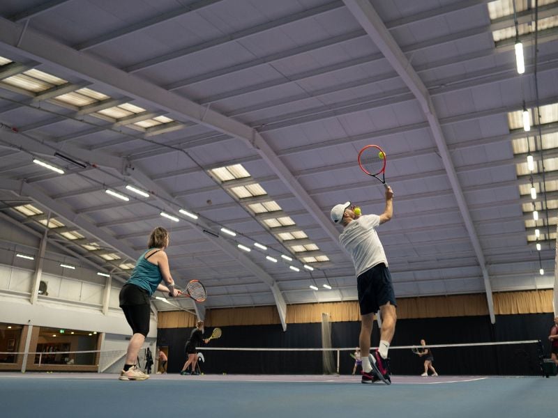 Indoor tennis game at DL Finchley, featuring multiple players with one serving on a well-lit court.