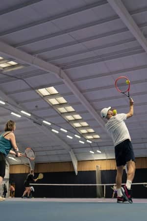 Indoor tennis game at DL Finchley, featuring multiple players with one serving on a well-lit court.