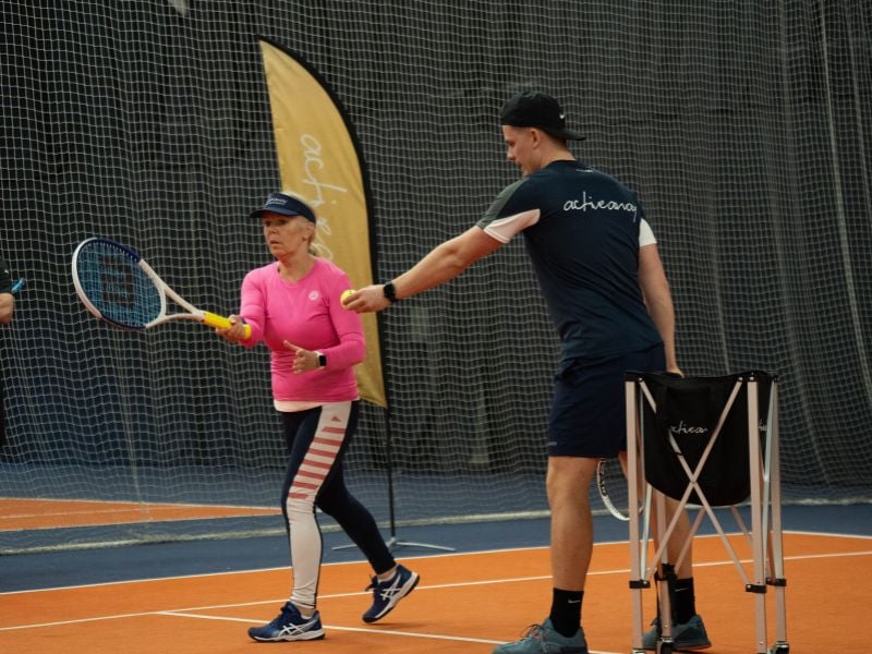 Senior woman receiving tennis coaching from a young male trainer in a blue 'active' outfit on an indoor court at DL Hull.