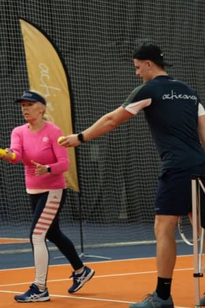 Senior woman receiving tennis coaching from a young male trainer in a blue 'active' outfit on an indoor court at DL Hull.