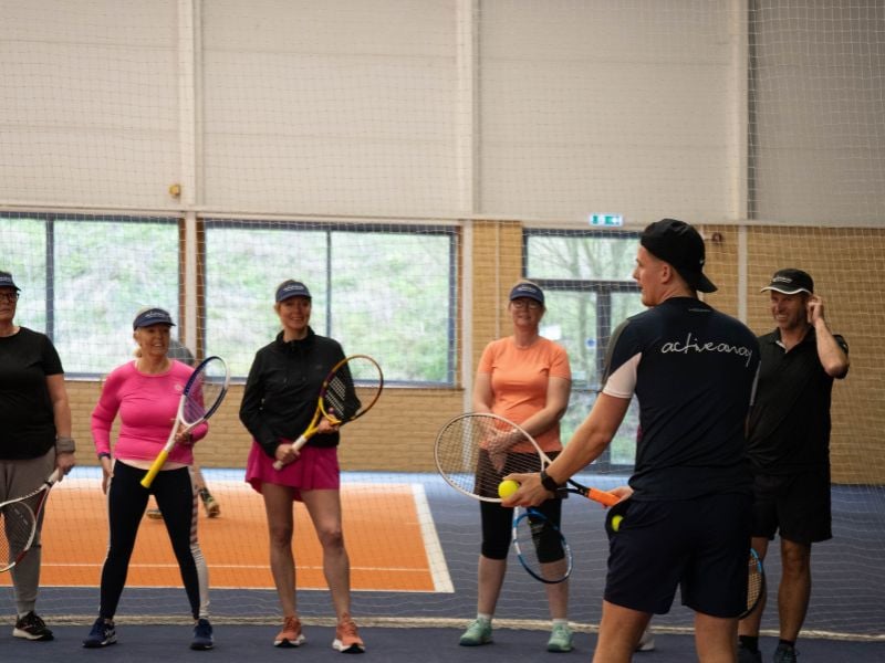 Tennis group coaching session at DL Hull with coach Alex instructing five players holding rackets on an indoor court.