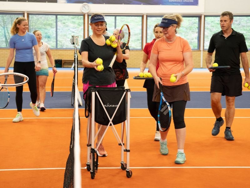 Tennis players at DL Hull Gallery preparing for drills, holding rackets and balls on a vibrant orange and blue court.