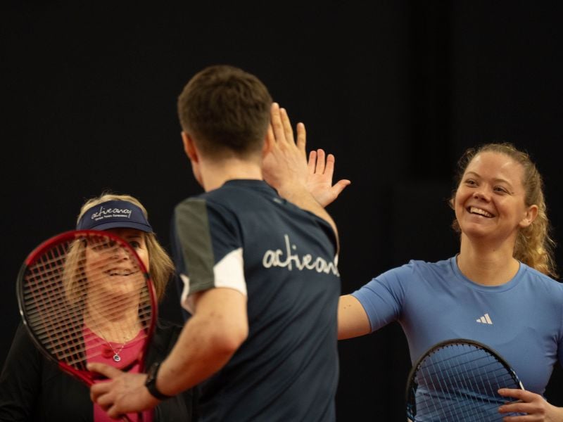 Tennis players sharing a high five at DL Hull Gallery, with a smiling onlooker nearby.