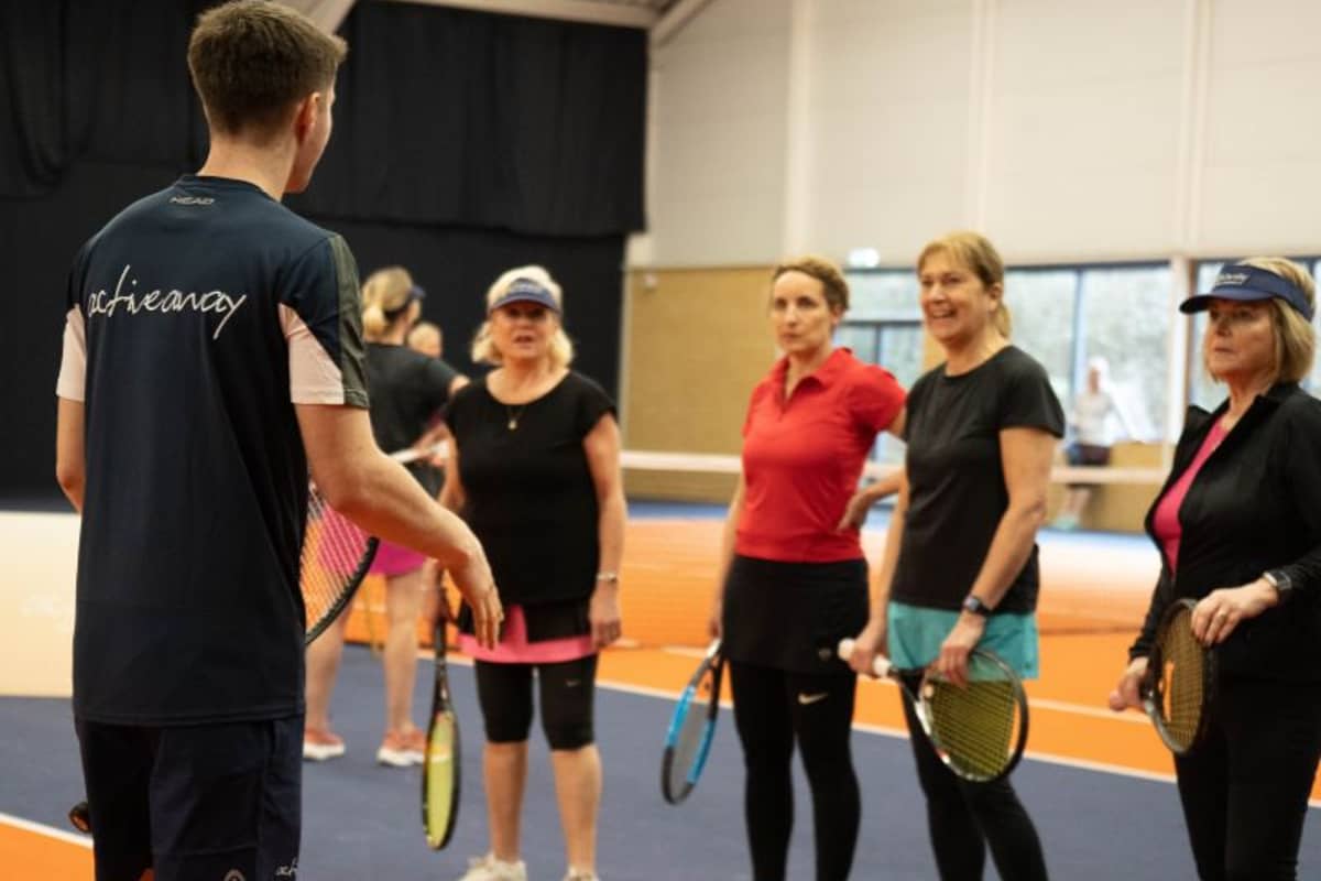 Tennis coach Jack at DL Hull instructs a group of female players on the indoor court, who are holding rackets and wearing sportswear.
