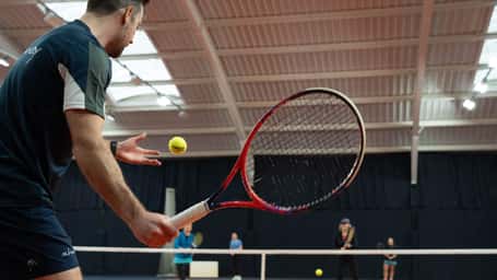 Indoor tennis coaching session with a coach preparing to serve, players in the background watching on a court with skylight windows.