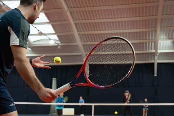 Indoor tennis coaching session with a coach preparing to serve, players in the background watching on a court with skylight windows.