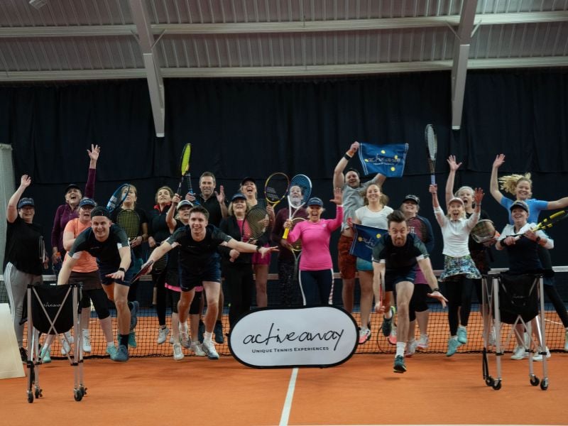 A group of excited tennis players jumping with rackets on an indoor court, promoting Active Away's unique tennis experiences.