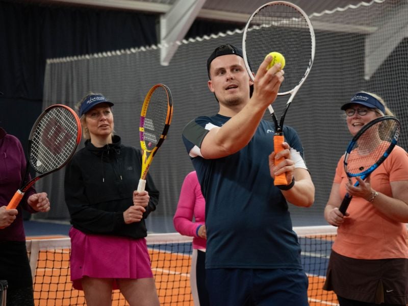Indoor tennis coaching session with coach Alex showing a technique to adult players standing on a clay court.