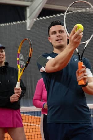 Indoor tennis coaching session with coach Alex showing a technique to adult players standing on a clay court.