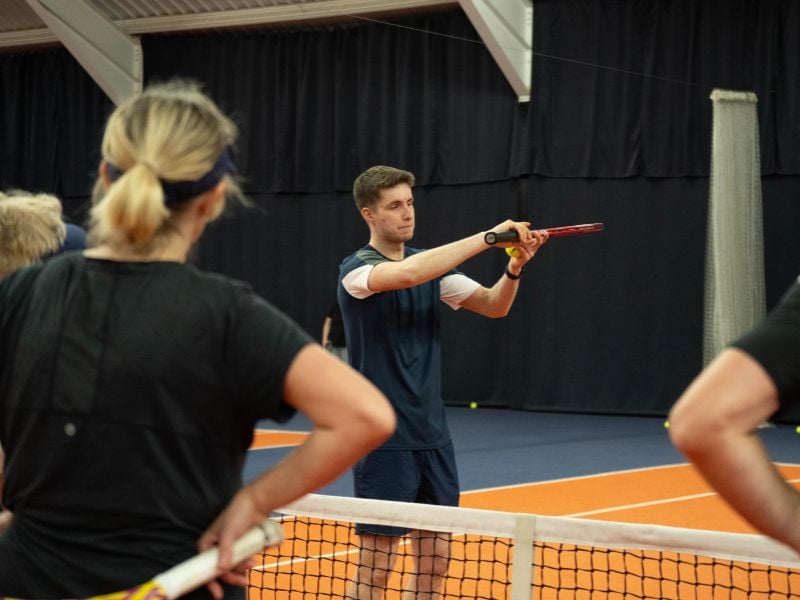 Tennis coach Jack demonstrates a forehand stroke to adult players at an indoor court in MK Gallery.
