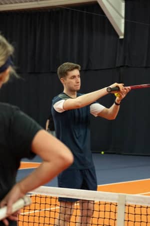 Tennis coach Jack demonstrates a forehand stroke to adult players at an indoor court in MK Gallery.
