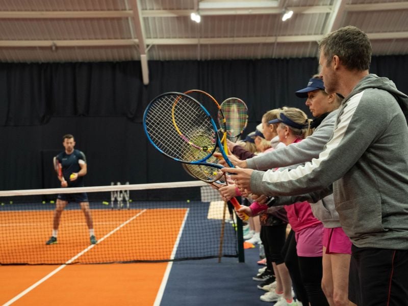 Group of people during a tennis coaching session with instructor Josh at DL MK Gallery, all holding racquets and standing on an indoor court.