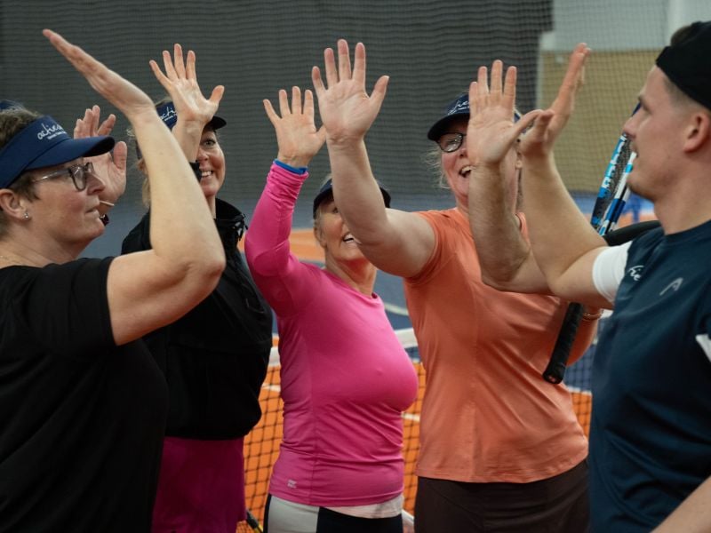 Five women in sports gear exchanging high fives on a tennis court, each holding a tennis racquet, showing teamwork and joy.