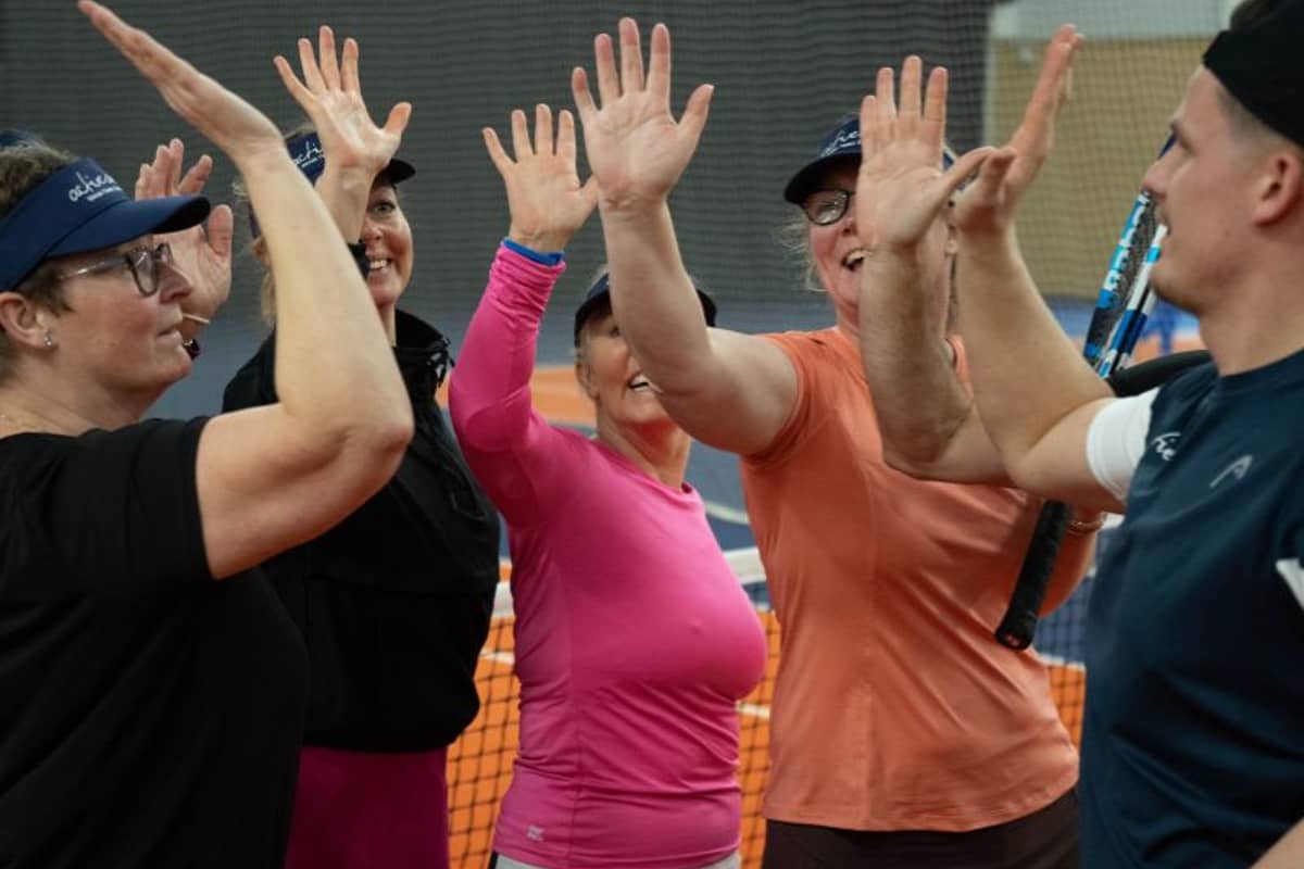 Five women in sports gear exchanging high fives on a tennis court, each holding a tennis racquet, showing teamwork and joy.