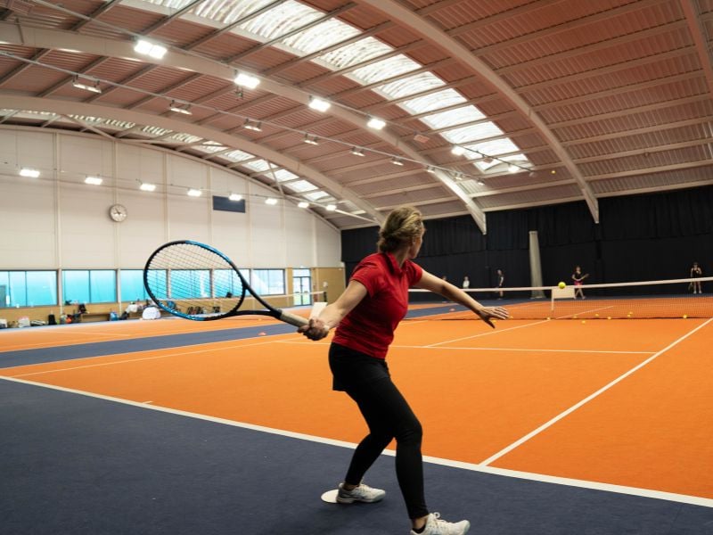 Woman playing tennis on an indoor orange court at David Lloyd Milton Keynes, preparing to return a shot with other players in the background.