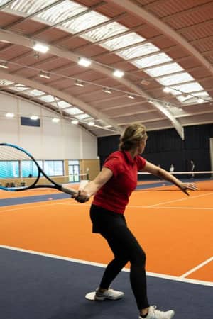 Woman playing tennis on an indoor orange court at David Lloyd Milton Keynes, preparing to return a shot with other players in the background.