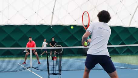 Indoor tennis doubles match with two teams competing under a green and white dome, players focused on returning the ball.