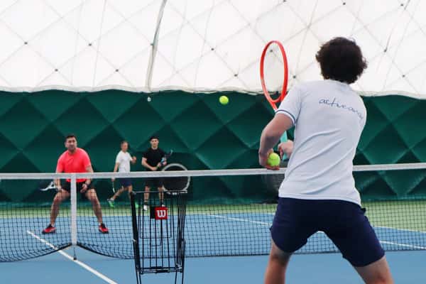 Indoor tennis doubles match with two teams competing under a green and white dome, players focused on returning the ball.