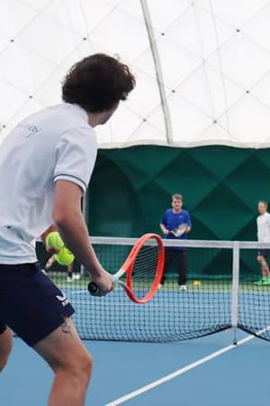 Group playing tennis indoors at DL Northwood Gallery, with a young man in white ready to hit the ball using an orange racquet.