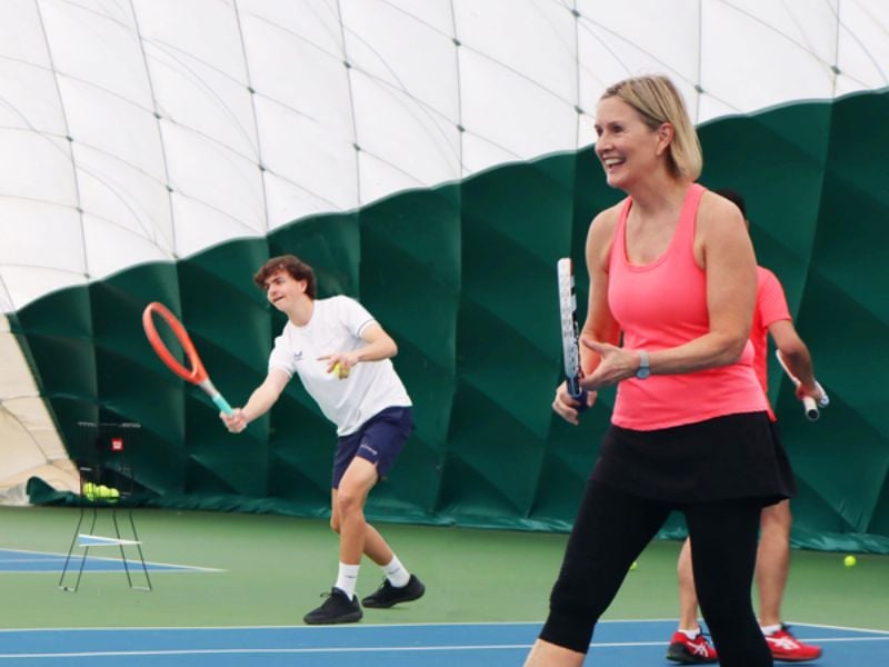 Indoor tennis lesson at DL Northwood with a coach smiling and a student preparing to hit a ball.