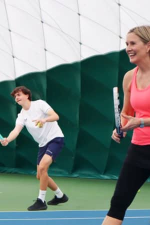 Indoor tennis lesson at DL Northwood with a coach smiling and a student preparing to hit a ball.